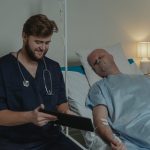Profissional de saúde consulta um tablet enquanto acompanha paciente hospitalizado em um leito.