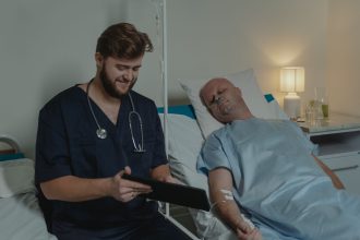 Profissional de saúde consulta um tablet enquanto acompanha paciente hospitalizado em um leito.