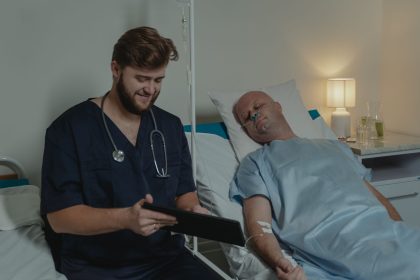 Profissional de saúde consulta um tablet enquanto acompanha paciente hospitalizado em um leito.