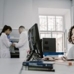 Mulher concentrada trabalha em um computador em um laboratório, com equipamentos e outras pessoas ao fundo.
