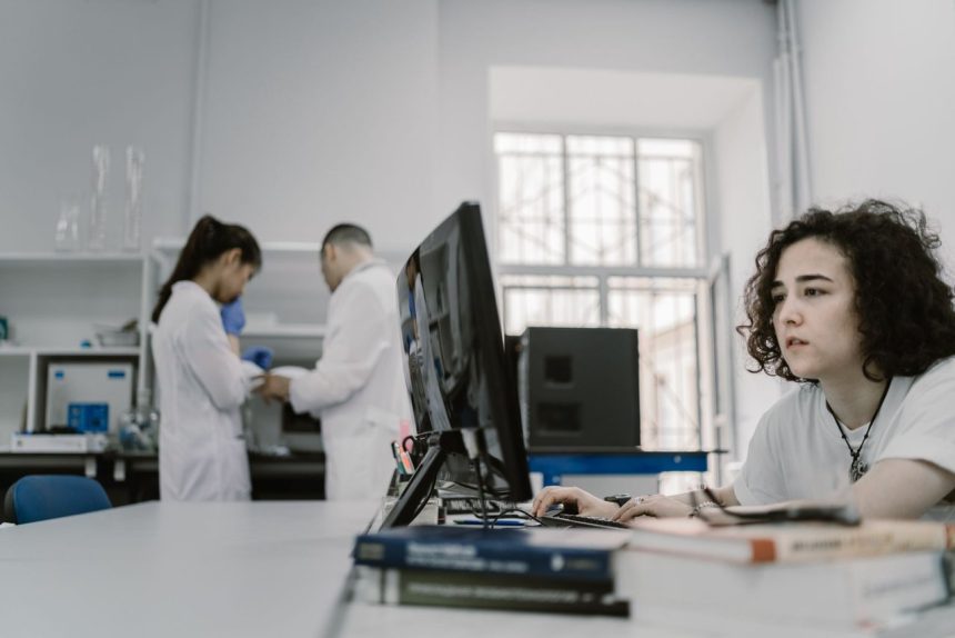 Mulher concentrada trabalha em um computador em um laboratório, com equipamentos e outras pessoas ao fundo.
