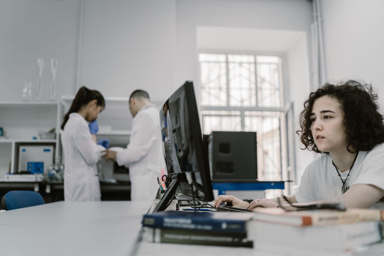 Mulher concentrada trabalha em um computador em um laboratório, com equipamentos e outras pessoas ao fundo.
