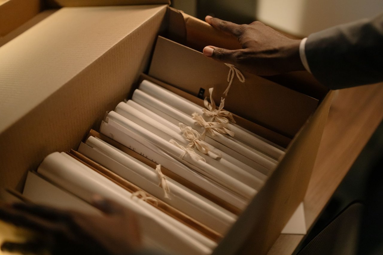 Mãos organizam documentos e pastas em uma caixa, em contexto de investigação.