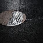 Tampa de bueiro molhada em uma rua urbana durante a chuva, refletindo a luz.
