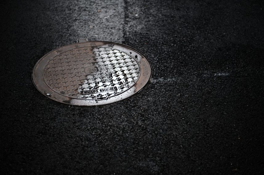 Tampa de bueiro molhada em uma rua urbana durante a chuva, refletindo a luz.